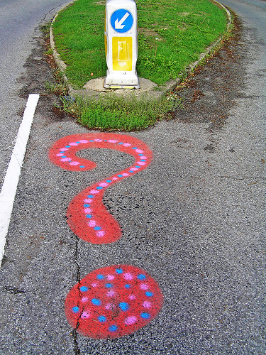 A red spray-painted question mark with blue and pink dots on tarmac, beside a traffic island with a keep-left bollard.