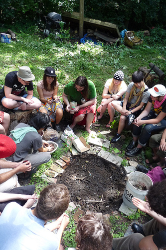 Photograph of a group of young people sitting in a circle on the ground outdoors, looking down at something in the middle.