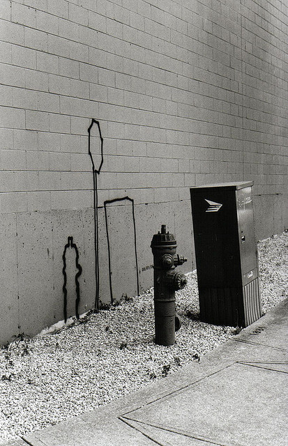 Black-and-white photograph of a fire hydrant and a metal utility box standing against a plain wall, with a hand-drawn outline on the wall behind them.