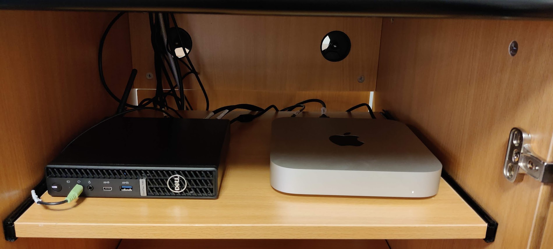 Photograph of a small Dell computer and a Mac mini side by side on a shelf in a wooden cabinet.