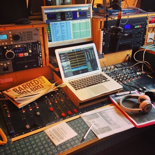Photograph of a radio studio desk with a laptop showing audio software, mixing desks and equipment racks, and a newspaper.