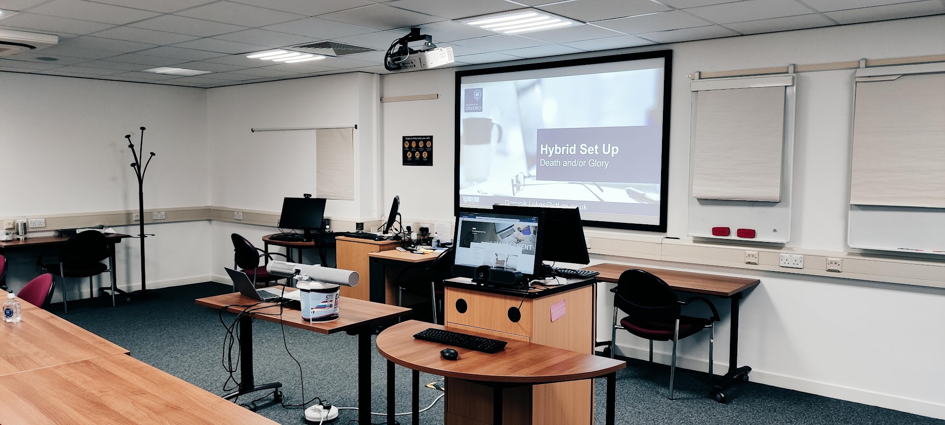 Photograph of the same seminar room from another side, with the projector screen showing “Hybrid Set Up”, the presenter's desk and four labelled arrows pointing at the equipment.