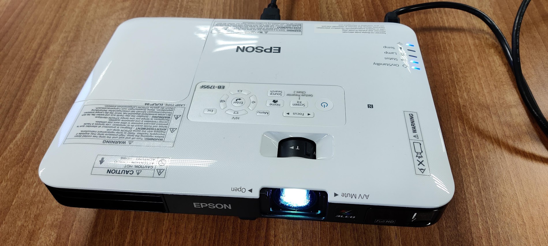 Photograph of a white Epson projector on a wooden desk, with its lamp lit.