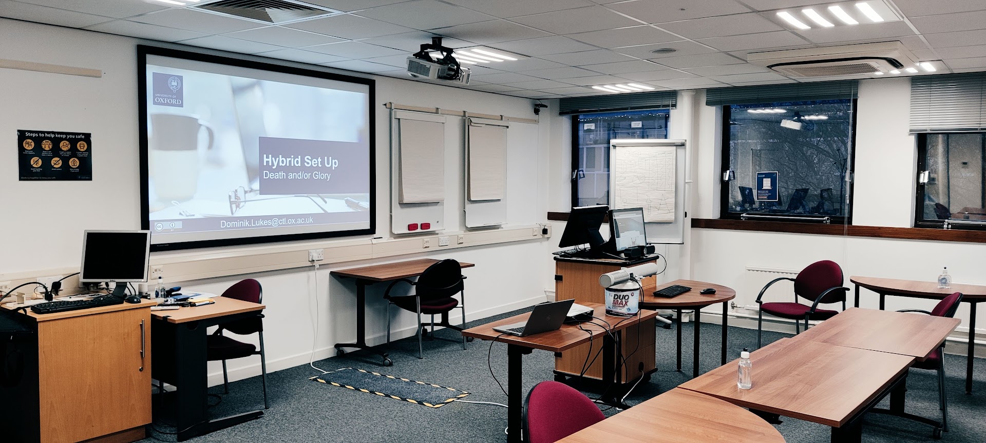 Photograph of a seminar room: a projector screen showing the slide “Hybrid Set Up”, desks with a laptop and a speaker-and-camera unit, and a media cabinet, with four labelled arrows pointing at the equipment.