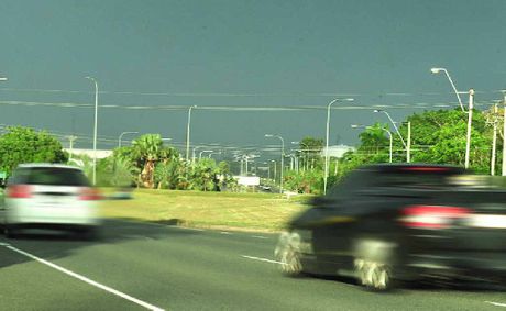 Photograph of cars on a road, blurred by their speed, with grass and trees beyond.