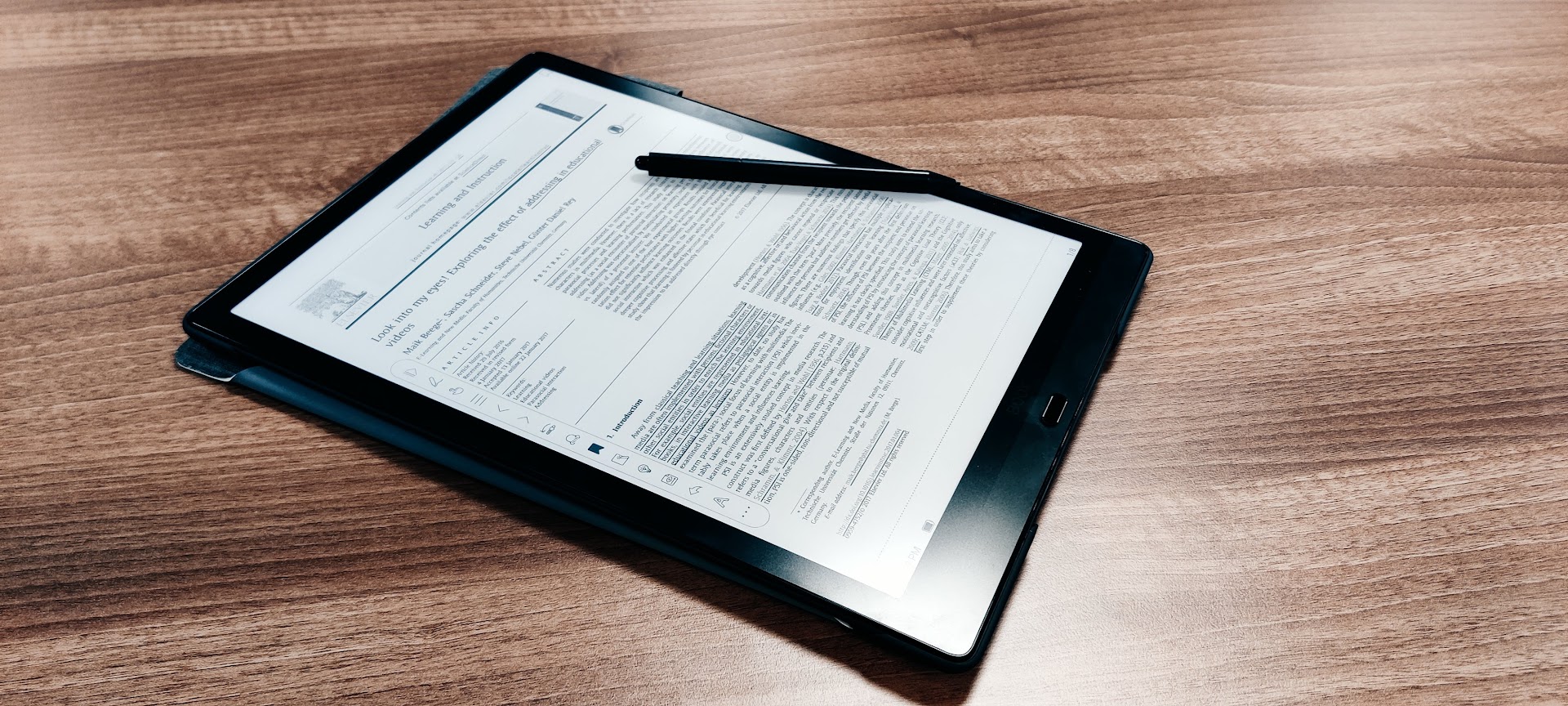 Photograph of a Boox Max Lumi e-ink tablet lying on a wooden table with a stylus.