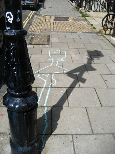 Photograph of a paved pavement with a black lamp post; a chalk outline of a lying figure, drawn in light blue, runs across the paving stones, with the lamp post’s shadow beside it.