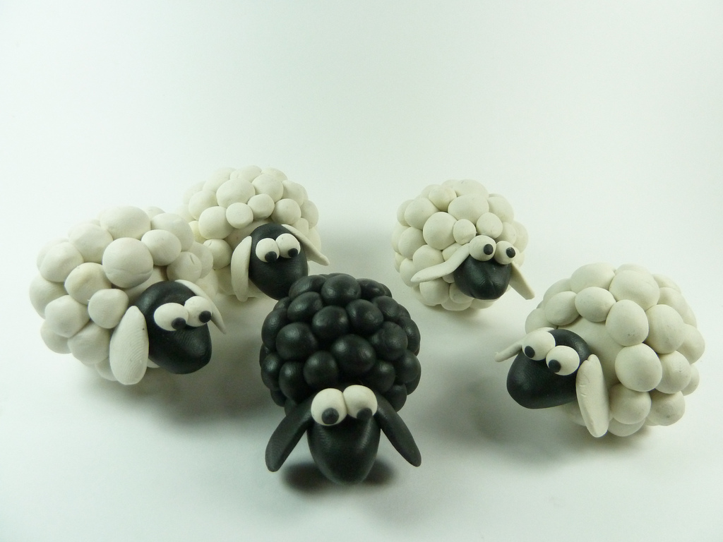 A photograph of small clay sheep: four white woolly sheep and one black sheep in the middle of them.