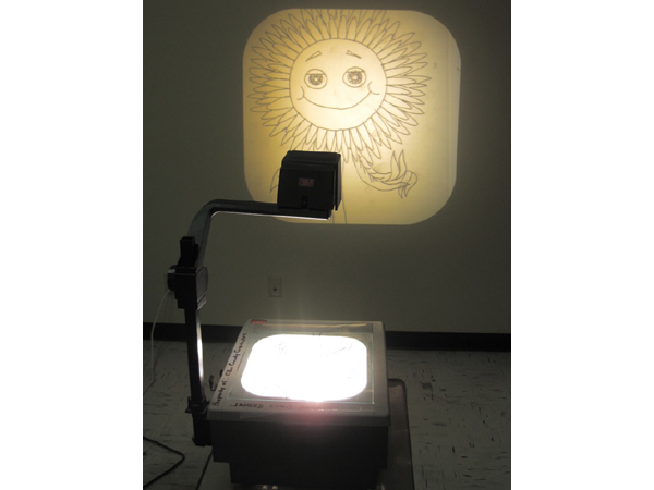 Photograph of an overhead projector in a dim room, projecting a drawing of a smiling sun onto the wall.