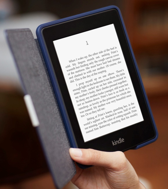 A hand holds a Kindle Paperwhite e-reader in a dark cover, showing a page of text.
