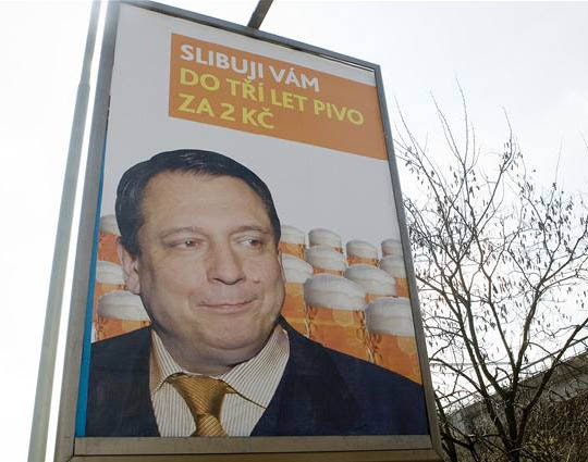 Photograph of a tall billboard seen from below against a bare tree: an orange banner with the text “Slibuji vám do tří let pivo za 2 Kč” above a close portrait of a man in a dark jacket and gold tie smiling to one side, with glasses of beer behind him.