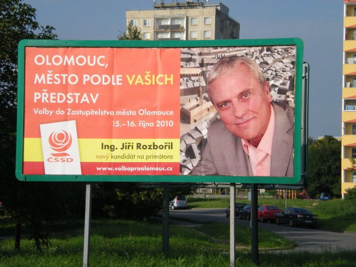 Photograph of an election billboard beside a block of flats: an orange and red panel reading “Olomouc, město podle vašich představ”, “Volby do Zastupitelstva města Olomouc 15.–16. října 2010”, a party logo, a web address, and a portrait of a smiling grey-haired man in a jacket with a caption naming him as a new candidate for mayor.