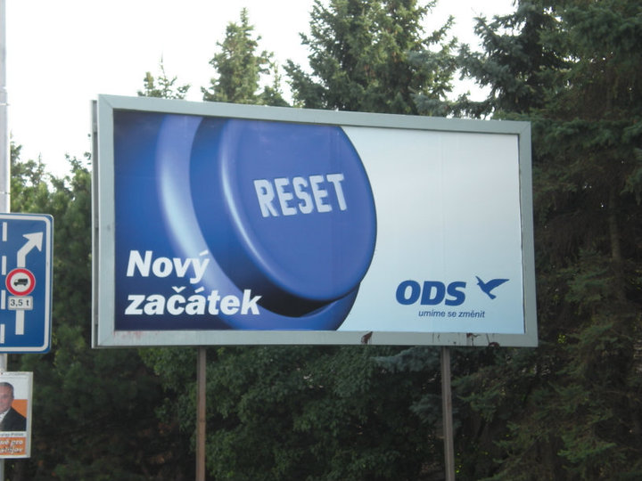Photograph of a billboard among trees showing a large blue “RESET” button with the text “Nový začátek” and the ODS logo with a short slogan.