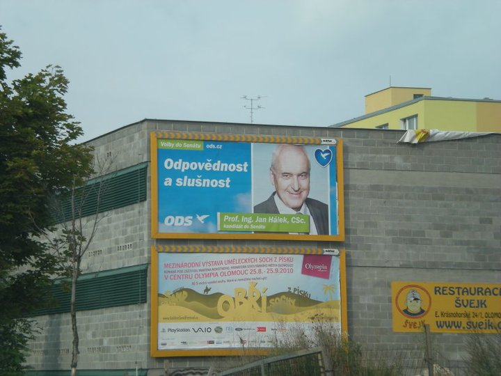 Photograph of a grey block wall carrying two yellow-framed billboards: the upper is a blue election advertisement with the text “Volby do Senátu”, “Odpovědnost a slušnost”, the ODS logo and a portrait of a white-haired man with a caption naming him as a Senate candidate; the lower advertises an international exhibition of sand sculptures at a shopping centre in Olomouc, 25.8.–25.9.2010.