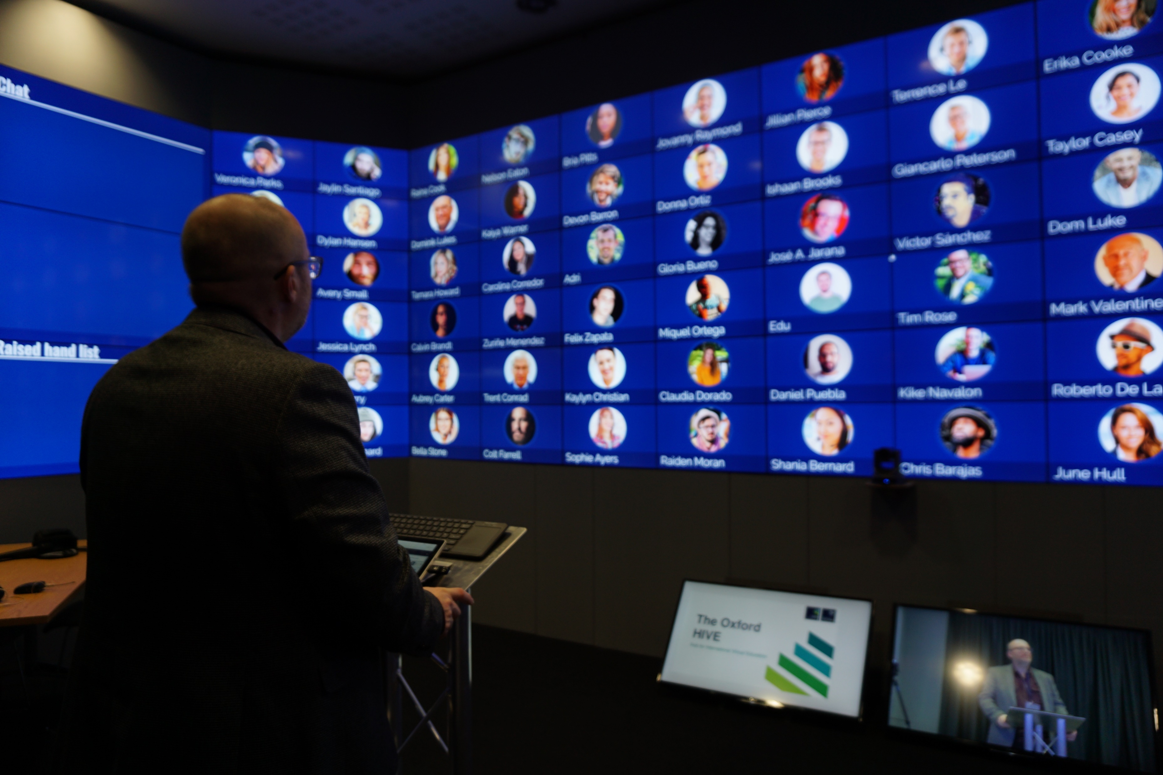 Photograph of a seated man seen from behind, looking at a wall of video-call participants, with a tracking camera in the foreground.