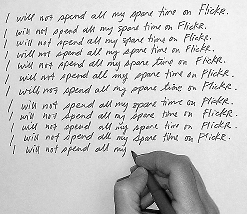 Black-and-white photograph of a hand writing “I will not spend all my spare time on Flickr.” repeatedly on a page of lined paper.