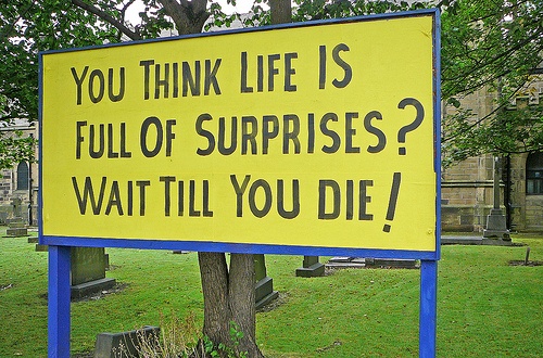 Photograph of a yellow roadside sign reading “You think life is full of surprises? Wait till you die!”